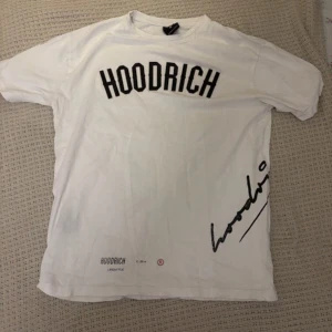 Vit HOODRICH t-shirt med shorts  - Vit t-shirt från HOODRICH med svart tryck framtill och textdetaljer på både fram- och baksida. Oversized vibe och cool streetstil, perfekt för dig som gillar statement-plagg. Materialet känns som bomull och t-shirten har klassisk passform med korta ärmar.