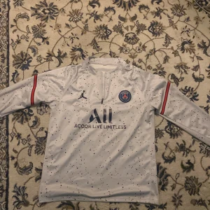 PSG långärmad vit fotbollströja Jordan - Snygg långärmad PSG-tröja i vitt med svarta prickar, broderat klubbmärke på bröstet och Jordan-logga. Röd och blå detalj på ärmarna, halv dragkedja framtill och tryck på ryggen. Tillverkad i lätt och ventilerande material, perfekt för träning eller match.