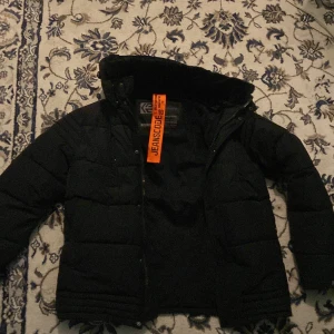 Svart pufferjacka från Jeanscode, S - Svart pufferjacka från Jeanscode med huva och diskret logga på ärmen. Jackan har quiltad design, dragkedja och knappar framtill. Perfekt för kalla dagar och har en modern, stilren look. Fodrad insida för extra värme.