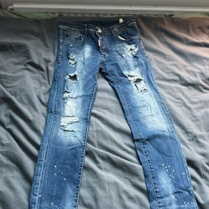 Dsquared2 blå jeans med slitningar W32 - Säljer ett par blå jeans från Dsquared2 i storlek W32. Byxorna har coola slitningar och hål på framsidan, samt vita färgstänk för en edgy look. Klassisk femficksmodell med knapp och dragkedja. Passar dig som gillar streetstyle och detaljer.