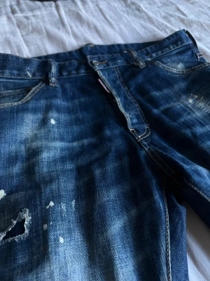 Dsquared2 Bros blå skinny jeans - Snygga blå jeans från Dsquared2 Bros med slitna detaljer, hål och färgstänk för en cool, edgy look. Skinny fit och klassisk femficksdesign, med brunt läderpatch bak och Dsquared2-logga vid gylfen. Perfekta för dig som gillar streetstyle.