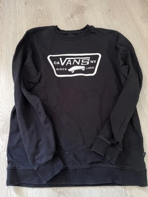 Svart Vans sweatshirt med tryck - Svart sweatshirt från Vans i storlek medium med stort vitt tryck framtill och skateboardmotiv. Tröjan har rund hals, ribbade muddar vid ärmslut och nederkant, samt långärmad passform. Perfekt för dig som gillar streetwear och skatekänsla.