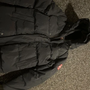Svart pufferjacka från Canada Goose - Säljer en svart pufferjacka från Canada Goose med huva och klassisk logga på ärmen. Jackan har flera stora fickor, dragkedja och tryckknappar framtill. Perfekt för kalla dagar och riktigt snygg streetstil.