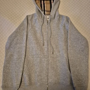 Grå hoodie från Burberry med dragkedja - Snygg grå hoodie från Burberry i storlek M med klassiskt rutigt foder i huvan. Tröjan har snörning vid huvan, praktiska fickor och hel dragkedja framtill. Perfekt för dig som gillar stilrena och exklusiva märken.