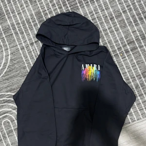 Svart AMIRI hoodie med regnbågstryck - Svart hoodie från AMIRI i 100% bomull med stor huva och färgglatt regnbågstryck både fram och bak. Snyggt AMIRI-märke i metall vid nederkanten och coolt drip-mönster i flera färger. Perfekt för dig som gillar streetwear och vill sticka ut.