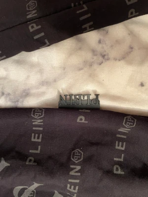 Svart långärmad tröja från Philipp Plein - Svart långärmad tröja från Philipp Plein med all-over logoprint i grått. Tröjan har rund hals och ribbade muddar. Snygg metallisk Plein-detalj vid nederkanten som ger en exklusiv vibe. Perfekt för dig som gillar streetwear och designer.