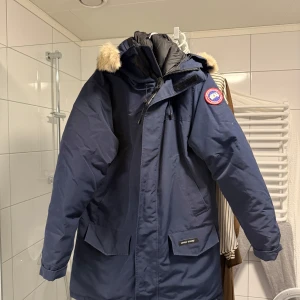 Mörkblå Canada Goose Langford parka - Säljer en mörkblå parkas från Canada Goose med ikonisk patch på ärmen och pälsdetalj på huvan. Jackan har stora fickor framtill, justerbar huva och klassisk rak passform. Perfekt för kalla vinterdagar och riktigt snygg till streetwear.