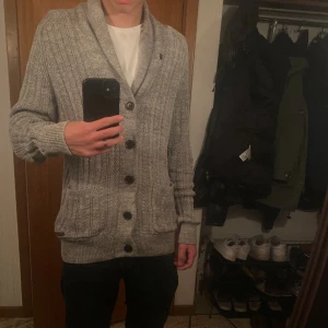 Grå ribbad kofta från Pier One - Säljer en grå ribbstickad kofta från Pier One med knappar och två fickor framtill. Koftan har bruna mockadetaljer på armbågarna och en bred krage som ger en chill vibe. Perfekt för lager-på-lager under hösten och vintern.