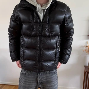 Svart pufferjacka från C.P. Company - Säljer en svart pufferjacka från C.P. Company med ikoniska goggles på huvan. Jackan har dragkedja framtill, två fickor och en glansig finish. Perfekt för kalla dagar och ger en riktigt clean streetwear-look.