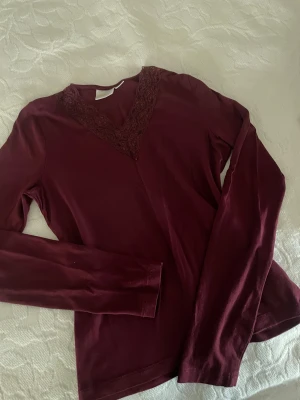 Vinröd långärmad tröja med spets - Snygg vinröd långärmad tröja med v-ringad hals och dekorativ spetsdetalj runt ringningen. Mjuk och skön bomullskänsla, perfekt för dig som gillar stilrena plagg med en liten twist. Passar dig som vill ha en enkel men ändå lite extra topp.