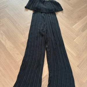 Svart plisserad byxdress från ONLY - Säljer en svart plisserad byxdress från ONLY i storlek 38. Byxdressen har breda ben, axelband och ett snyggt volangparti upptill. Materialet är lätt och ger ett flowigt intryck, perfekt för dig som gillar stilrena och trendiga plagg.