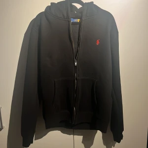 Svart hoodie från Ralph Lauren - Svart hoodie från Ralph Lauren med röd broderad logga på bröstet. Large men passar Medium också. Tröjan har huva, dragkedja framtill och två stora fickor. Tillverkad i 100% bomull för skön känsla. Perfekt för chill dagar och streetwear-stil.