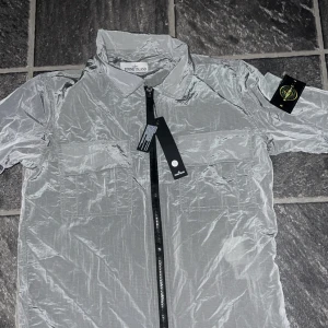 Silver vindjacka från Stone Island - Snygg och lätt silverfärgad vindjacka från Stone Island med klassisk logga på ärmen. Jackan har två bröstfickor med lock, hel dragkedja framtill och en glansig finish. Perfekt för dig som vill sticka ut med en modern och sportig look.