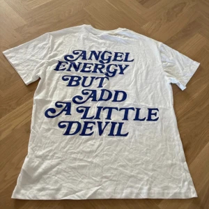 Vit t-shirt med blå text från FB Sisteroverzied t shirt - Vit t-shirt från FB Sister i storlek XXS med coolt blått tryck både fram och bak: 'Angel energy but add a little devil'. Klassisk rund hals och korta ärmar. Mjuk bomullskvalitet och relaxed passform, perfekt för dig som gillar statement-plagg.overzied t shirt.