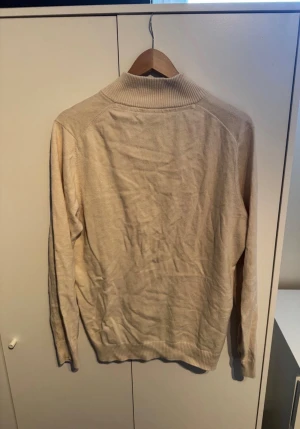 Half zip - Snygg beige långärmad tröja från Lyle & Scott med half zip och ribbad krage. Tröjan har det klassiska gula fågel-loggan broderad på bröstet och är tillverkad i mjuk bomull. Perfekt för dig som gillar stilrena och bekväma plagg.