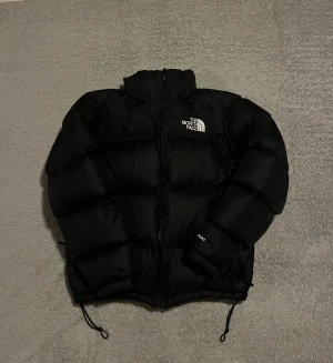 North Face Retro Nupste  - Säljer en svart dunjacka från North Face (Retro Nupste) Jackan har dragkedja framtill, hög krage och snörning vid nederkant. Perfekt för kalla dagar. 