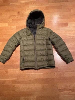 Olivgrön Peak Performance dunjacka - Snygg olivgrön dunjacka från Peak Performance med huva och svart foder. Jackan har quiltad design, dragkedja framtill och broderad logga på bröstet. Perfekt för kalla dagar.