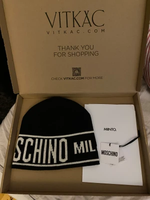 Svart och vit mössa från Moschino - Snygg svart mössa från Moschino med vit logotyptext runt om. Klassisk form och stilren design som sticker ut med den kontrasterande vita texten. Perfekt accessoar för att lyfta din vinterlook.