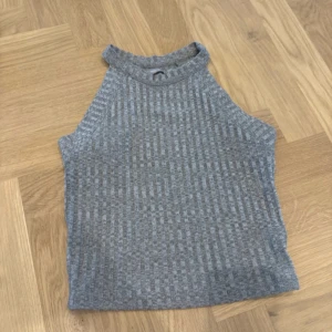 Grå ribbad linnetopp från Bershka - Säljer en grå ribbad linnetopp från Bershka med hög hals och ärmlös design. Toppen har ett snyggt vertikalt randigt mönster och är gjord i mjukt stickat material som känns skönt mot huden. Perfekt att styla med jeans eller kjol för en chill look.