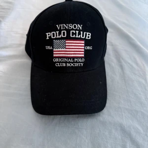 Svart keps Vinson Polo Club - Svart keps från Vinson Polo Club med böjd skärm och broderad amerikansk flagga framtill. Texten 'Original Polo Club Society' och 'USA' är broderad i vitt. Justerbar rem baktill. Klassisk och sportig stil, perfekt för dig som gillar preppy vibes.