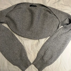 Grå ribbad bolero med långa ärmar - Säljer en grå stickad bolero med ribbad struktur och långa ärmar. Perfekt att slänga över en t-shirt eller linne för extra värme och stil. Enkel att matcha och ger en chill vibe till din outfit.