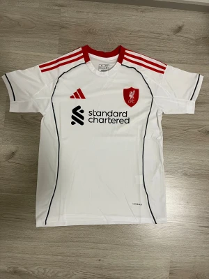 Vit Liverpool 25/26 borta fotbollströja Adidas M - Liverpool 25/26 borta fotbollströja i vitt med röda detaljer och svarta linjer. Adidas-logga och klubbmärke på bröstet, samt Standard Chartered-tryck. Tillverkad i lätt och ventilerande material (AEROREADY). Klassisk passform med korta ärmar och rund hals.