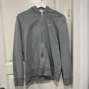 Grå hoodie från Nike med dragkedja - Snygg grå hoodie från Nike med vit broderad logga på bröstet. Tröjan har huva med snörning, praktiska fickor framtill och hel dragkedja. Perfekt för chill eller träning, tillverkad i mjuk bomullsmix för skön komfort.