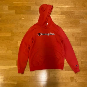Röd Champion hoodie med huva - Riktigt snygg röd hoodie från Champion med stor logga broderad på bröstet och en mindre logga på ärmen. Tröjan har huva med snörning och en stor magficka framtill. Mjuk och skön i bomullsmix, perfekt för chill eller streetwear.