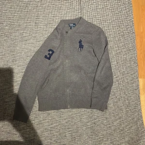 Grå stickad cardigan från Polo Ralph Lauren - Snygg grå stickad kofta från Polo Ralph Lauren med dragkedja framtill, mörkblå broderad polospelare på bröstet och en stor mörkblå siffra 3 på ärmen. Ribbad nederkant och muddar, perfekt för chilliga dagar.
