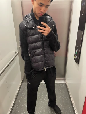 Svart dunväst från Moncler med huva - Säljer en svart dunväst från Moncler med huva och dragkedja framtill. Västen har quiltad design och Moncler-logga på vänster ärm. Perfekt för lager-på-lager och har en modern, loose passform. Grym att styla med både jeans och mjukis. Dm för rabbat 