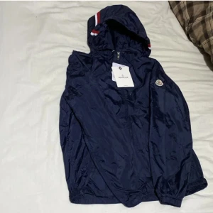 Mörkblå vindjacka från Moncler - Snygg mörkblå vindjacka från Moncler med huva och dragkedja. Jackan har Moncler-logga på ärmen och rödvit rand på huvan. Tillverkad i glansig polyester, perfekt för vår och höst. Klassisk passform och elastiska muddar vid ärmslut och midja.