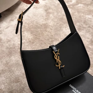 Svart axelväska från Saint Laurent - Snygg svart axelväska från Saint Laurent Paris i slät skinn med justerbar rem och ikonisk YSL-logga i guld på framsidan. Väskan har en stilren, rektangulär form och dragkedja på insidan. Perfekt för dig som gillar lyxiga detaljer och klassisk design.