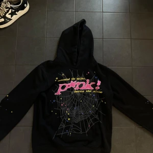 Spider hoodie  - Svart spider hoodie med stor huva och tryck på bröstet med texten 'Pink!' och spindelnätsmotiv. Små stjärnor i gult, rosa och blått över hela framsidan. På ryggen finns en gul stjärna. Perfekt för dig som gillar streetwear och unika prints.