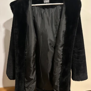 Svart pälsjacka med huva Urban Classics - Mysig svart pälsjacka från Urban Classics med stor huva och lång ärm. Jackan är fluffig och har en enkel, rak passform. Perfekt för kalla dagar när du vill vara både varm och snygg. Har använt den totalt två gånger.