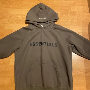 Grå Essentials hoodie med huva - Säljer en grå hoodie från Essentials med stor svart logga på bröstet. Tröjan har huva, känguruficka och långa ärmar. Mjuk och skön bomullsmix som passar perfekt för chill eller streetwear. Oversized passform för en avslappnad vibe.