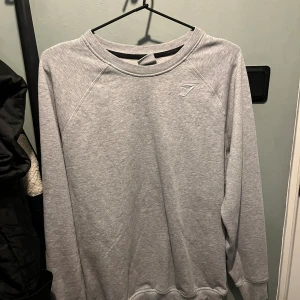 Grå sweatshirt från Gymshark - Snygg grå sweatshirt från Gymshark i storlek S. Tröjan har rund hals, ribbade muddar och ett diskret Gymshark-logo broderat på bröstet. Perfekt för chill eller träning, med långärmad passform och mjukt material.