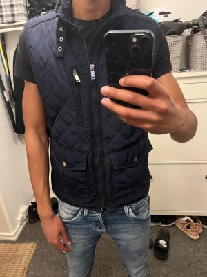 Väst - Snygg mörkblå quiltad dunväst med hög krage och silverfärgade detaljer. Västen har två stora fickor med tryckknappar, en bröstficka med dragkedja och coola metallöljetter vid halsen. Perfekt att slänga över en hoodie eller t-shirt för extra stil.