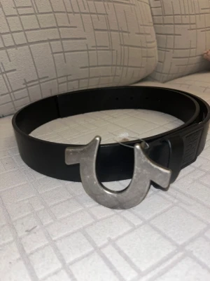 Svart True Religion skärp med hästskospänne - Säljer ett svart skärp från True Religion i äkta skinn med ett rejält metallspänne format som en hästsko. Bältet har en stilren och clean look, perfekt för att lyfta jeans eller byxor. Märkets logga är präglad på lädret nära spännet.