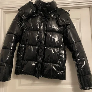 Svart pufferjacka Calvin Klein Jeans - Säljer en svart, glansig pufferjacka från Calvin Klein Jeans med ikoniskt CK-logo på bröstet. Jackan har huva, dragkedja. Säljer pga för liten. Köpt för 2199kr