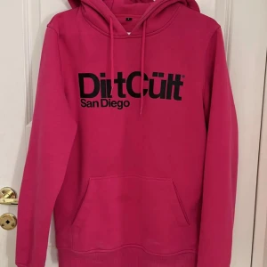 Rosa hoodie från DirtCult San Diego - Säljer en snygg rosa hoodie från DirtCult San Diego med svart tryck på bröstet. Hoodien har huva med snörning och en stor magficka framtill. Perfekt för dig som gillar streetwear och vill sticka ut med färg.