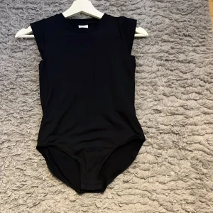 Svart ärmlös bodysuit - Säljer en svart bodysuit med rund hals och breda axelpartier. Toppen är helt slät utan mönster och har en tight passform som framhäver siluetten. Materialet känns mjukt och stretchigt, perfekt för att styla med jeans eller kjol.