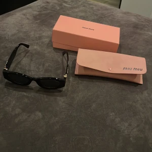 Svarta Miu Miu solglasögon med guld - Snygga svarta solglasögon från Miu Miu med rektangulär form och coola detaljer i guld på skalmarna. Glasögonen är tillverkade i Italien och kommer med original rosa fodral och ask. Perfekt accessoar för att lyfta din stil.