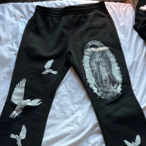 Svarta mjukisbyxor med tryck - Svarta mjukisbyxor med elastisk midja och coola vita tryck av duvor samt en stor bild av Jungfru Maria på ena benet. Byxorna har sidofickor och är gjorda i mjukt material som känns skönt mot huden. Perfekt för dig som gillar streetwear med unik design.