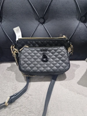 Svart quiltad axelväska från Guess - Snygg svart axelväska från Guess med quiltat mönster och guldfärgade detaljer. Väskan har två fack med dragkedja, kedja och justerbar axelrem. Guess-logga och G-emblem framtill ger en cool touch. Perfekt storlek för det viktigaste.