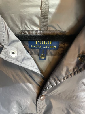 Grå vindjacka från Polo Ralph Lauren - Snygg grå vindjacka från Polo Ralph Lauren med klassisk krage och knäppning framtill. Jackan har diskret broderad logga på bröstet och två sidofickor. Tillverkad i glansig polyester som ger en clean och modern look.