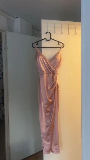 Balklänning  - Snygg långklänning i champagnefärgad satin med omlott-design och smala axelband. Med slits framtill. Klänningen har en v-ringad front och ett elegant fall som ger en lyxig känsla. Perfekt för dig som vill sticka ut med en stilren och modern look. Storleken kan justeras men är främst för xss-xs och s💕