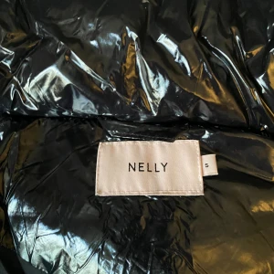 Svart glansig pufferjacka från Nelly - Svart pufferjacka från Nelly med glansig finish och huva. Jackan har dragkedja framtill och två fickor. Endast testad så mycket fint skick. 
