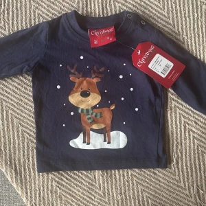 Mörkblå långärmad tröja med renmotiv till baby  - Till baby Mörkblå långärmad tröja från Christmas Edition i mjuk bomull. Framsidan har ett gulligt tryck med en ren som står på snö och vita prickar som snöflingor. Rund hals med tryckknappar på axeln för enkel påklädning. Perfekt för julens mysiga dagar.