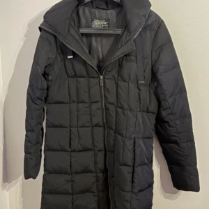 Svart lång pufferjacka från Tenson - Svart lång pufferjacka från Tenson med quiltad design och huva. Jackan har dragkedja framtill och raka ärmar. Perfekt för kalla dagar och har en stilren look som funkar till det mesta.fickan kedja är trasig 🌸