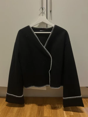 Svart omlottopp från Gina Tricot - Svart omlottopp från Gina Tricot med vita dekorativa sömmar längs kanterna. Toppen har lång ärm och croppad passform, vilket ger en modern och cool look. Perfekt för dig som gillar stilrena detaljer och kontraster.